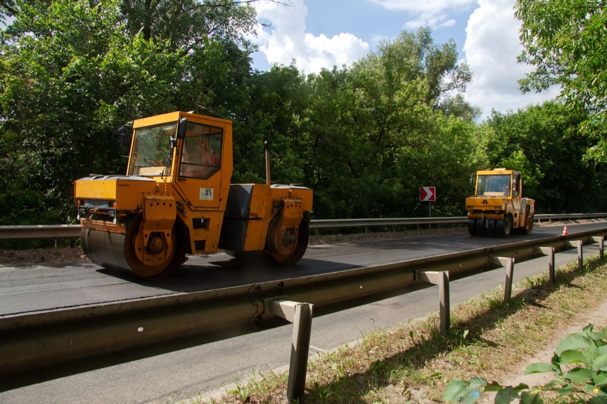 Two tandem rollers compacting a freshly laid asphalt carriageway on a tree-lined road